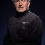 An elderly man with short gray hair is facing the camera. He is wearing a black Reebok jacket with logos on the chest. The background is dark, highlighting the subject's serious facial expression.