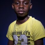 A young boy wearing a bright yellow "New York City 23" T-shirt sits against a dark blue background. The lighting highlights his facial features and the details of his T-shirt. He has a calm, neutral expression.