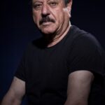 A man with short, dark hair and a salt-and-pepper mustache is wearing a black t-shirt. He has a serious expression and is set against a dark, plain background. The lighting creates shadows, emphasizing the contours of his face and arms.