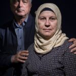 A middle-aged man and woman are posing against a dark background. The man, with gray hair, is standing and has his hand gently resting on the woman's shoulder. The woman, who is sitting, is wearing a patterned top and a beige hijab, looking directly at the camera.