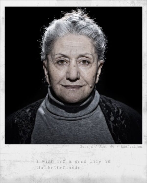 A black-and-white portrait of an older woman with gray hair pulled back, wearing a dark turtleneck and dark outer garment. Text below reads: "Suraja / Age: 69 / Azerbaijan. I wish for a good life in the Netherlands.