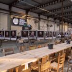 A spacious industrial gallery features portraits hanging along one wall below a "Kunst Reserve Bank" sign. In the center, a long wooden table with multiple chairs and potted plants creates an inviting communal area, illuminated by natural light from large windows.