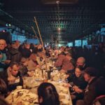 A crowded indoor gathering where people of various ages are seated and standing at long tables, eating and socializing. The dim lighting creates a cozy atmosphere, with conversations and shared meals giving the scene a festive and communal feel.