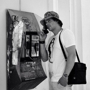 A man wearing a hat and a T-shirt with a woman's face on it is using a payphone. The scene, captured by photographer Rick Akkerman, shows him with a shoulder bag and a watch on his left wrist, standing next to a worn-out, graffitied payphone attached to the wall.