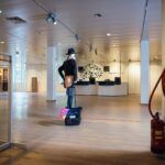 A spacious room with a wooden floor and ceiling lights illuminating the area. There is a mannequin dressed in casual clothing and a hat, standing on a black crate. A large tree decal and the words “Stop right at zal” are visible on the back wall near a reception desk.