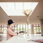 A fashionably dressed mannequin wearing a black hat and scarf sits at a white, modern counter with a coffee cup and saucer. A large flower in a vase is placed in front of the mannequin. The spacious room has large windows and a bright skylight above.