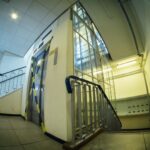 A person wearing a hat and carrying a bag stands at the bottom of a small set of stairs next to an elevator with caution tape. The hallway is lit with fluorescent lights and has industrial elements like pipes and a metal railing.