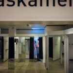 A fitting room area in a store with multiple booth-style rooms. The rooms have mirrors and black curtains for privacy. Two people are visible in the background, inside a fitting room. The partially visible sign above reads "paskamer.