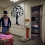 A mannequin dressed in casual clothing and a hat stands near a wall adorned with a black tree illustration. A kiosk and scale are positioned nearby. To the right, an open doorway leads to a restroom. In the foreground, a table with a pink checkered cloth and a chair.