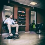 A man with a friendly smile sits in a barber chair inside a modern barbershop. The shop has warm colors, large mirrors, neatly arranged hair products on shelves, and another barber chair in the background. The space has a clean, inviting atmosphere.