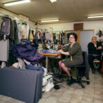 A woman sits at a sewing machine in a cluttered tailor shop, wearing a green dress and a measuring tape around her neck. A man sits in the background, smiling. The shop is filled with hanging clothes, sewing materials, and various tools.