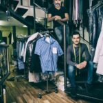 Two men in a clothing store, surrounded by clothes on racks. One man stands on a staircase, while the other sits on the bottom step, wearing a measuring tape around his neck. The store has wooden floors and various hanging garments.