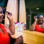 A woman in a red sleeveless top, smiling and holding a blue comb near her hair, looks at her reflection in a large mirror. Various hair care products are placed on the wooden counter in front of the mirror, and the background suggests a cozy salon setting.