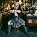 A man with white hair and beard sits confidently on a chair inside a cozy, dimly lit pub, wearing a plaid kilt and black shirt. His hands rest on a cane. The pub is decorated with various memorabilia, wooden furnishings, and low-hanging lights.