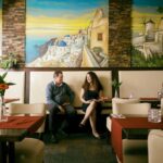 Two people sit and chat in a warmly lit restaurant with beige chairs and red tablecloths. The walls behind them feature large, colorful murals depicting a scenic coastal village, giving the space a vibrant and inviting atmosphere.