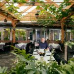 A man dressed in a dark shirt sits alone at a table in a well-lit, spacious restaurant with wooden beams and a lush, green vine canopy. Surrounding him are tables with white checkered tablecloths, and the ambiance is cozy and inviting.