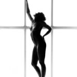 A silhouetted figure of a nude person standing gracefully on one leg with the other leg slightly bent. One hand is resting on the hip, while the other reaches up towards a tall vertical window behind them. The background is bright, highlighting their form.