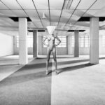 A surreal black and white image of a person standing in the center of a spacious, empty room with a grid-like ceiling. The person is nude, wearing high heels, and has a large cone-shaped object covering their head. The room is stark, with columns and large windows.