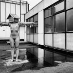 A nude person stands on a rooftop with arms outstretched, wearing a birdhouse on their head. The scene is reflected in puddles on the ground. The building in the background has large windows and a tiled exterior. The image is in black and white.