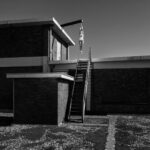 A black-and-white photo of a two-story brick building with a staircase leading to the upper level. A mannequin hangs from a suspended beam near the top of the stairs. The sky is clear, and the ground is covered with gravel.