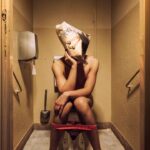 A person is sitting on a toilet with their underwear around their ankles. They have a paper bag over their head with a photo of another person's face printed on it. The restroom is narrow and dimly lit, with beige walls and a wall-mounted toilet paper dispenser.