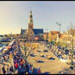 A bustling town square buzzes with activity during a daytime festival. Crowds gather near various stalls and a church tower rises prominently in the background. The scene is bathed in sunlight, casting a warm glow over the vibrant and festive atmosphere.