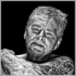 Black and white portrait of an elderly man with weathered skin and tattoos on his arm, gazing solemnly at the camera. His partially visible arm rests on the edge of the frame, and there is a high contrast between his face and the dark background.