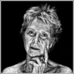 A grayscale portrait of an older person with short, tousled hair. They have a contemplative expression, gazing directly at the camera, with one hand resting against their chin and a prominent ring on their finger. The background is solid black.