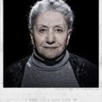 A portrait of an older woman with gray hair, wearing a dark shawl and a gray turtleneck. The lighting casts shadows behind her. Below the image, text reads: "Suraja / Age 69 / Azerbaijan. I wish for a good life in the Netherlands.