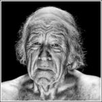 A black and white close-up portrait of an elderly man with a solemn expression. The man has a visibly wrinkled face, thinning white hair, and is shirtless against a dark background. The contrast highlights the texture and lines of his aged skin.