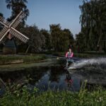 A person water skiing on a narrow canal at dusk, creating a large splash behind them. A traditional windmill and lush greenery line the canal banks, reflecting on the water under the evening sky. The person wears brightly colored gear and appears to be enjoying the activity.