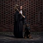 A person dressed in a judge's robe stands holding a large wooden gavel over their shoulder. They are accompanied by a large dog sitting at their side. The background is a brick wall with a distinct pattern. The lighting is moody and dramatic.