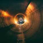 A glowing, cylindrical tunnel with warm orange and yellow light reflections, revealing a small circular opening at the far end. The lighting creates a mysterious and futuristic atmosphere, with sparks and particles visible near the center.