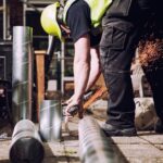 A construction worker wearing a yellow hard hat and high-visibility vest cuts a metal pipe with a power tool, creating a shower of sparks. Several spiral metal pipes lie on the ground nearby. The background includes construction materials and greenery.