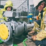 Two construction workers wearing safety gear, including helmets and gloves, work on installing or maintaining industrial piping machinery outdoors. One holds a large wrench, and the other monitors the process. Green straps secure the equipment, and a fence is visible in the background.