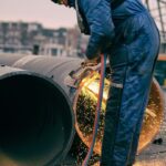 A worker in blue protective clothing and a beanie uses a cutting torch to cut through a large metal pipe. Sparks fly from the point of contact between the torch and the pipe. The background features industrial equipment and an overcast sky.