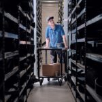 A person wearing a light blue shirt and cap stands between tall shelves full of various items in a dimly lit warehouse. They are leaning on a cart holding boxes and parts, looking slightly upward. The warehouse has a long aisle and industrial lighting overhead.