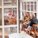 Two women are sitting at a wooden table in a bright, cozy café, engaged in conversation. One is wearing a yellow scarf, and they both have drinks in front of them. The café has large windows, letting in natural light, and colorful flowers are on a nearby table.