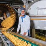 A worker wearing a hair net, face mask, and white uniform stands next to a conveyor belt in a factory. The belt is carrying a large quantity of chips from a cylindrical machine. The worker is smiling and the factory machinery surrounds him.