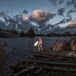 A person in a wetsuit holding a surfboard stands at the edge of a wooden dock by a large body of water, with dramatic clouds in the evening sky and houses visible in the background. Grasses and reeds grow by the dock.