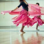 A woman gracefully twirls in a bright dance studio, her pink skirt flowing around her. She smiles, looking at the camera, with her reflection visible in the wall mirror behind her. The floor is made of polished wood, and the room is flooded with natural light.