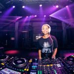 A DJ smiles while standing behind a professional DJ booth with multiple mixers and turntables. She wears headphones and a black shirt with a graphic design. Purple stage lights and a stylish background create a lively atmosphere.