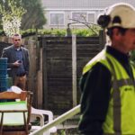 A man in a construction hard hat and high-visibility vest stands in the foreground while another man in a suit and patterned pants stands in the background near wooden gates. Various chairs and crates are scattered around the yard.