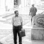 A man in a light shirt and dark pants holds a briefcase, standing on a sunlit street next to an ornamental stone object. Another person walks away in the background. The scene is in black and white.