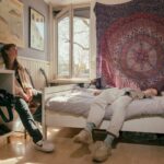 A cozy bedroom with two people relaxing. One person sits on a chair near a window, while the other lies on a bed with an intricate tapestry hanging behind it. The room features various decorations, shoes on a shelf, and soft natural light filtering through the open window.