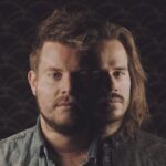 Two men with serious expressions stand closely together against a dark, patterned background. One has short hair and a beard, while the other has long hair and a beard. Subdued lighting casts shadows on their faces, partially obscuring their features.