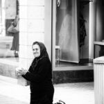 A black-and-white photo of a woman dressed in dark clothing and a headscarf, kneeling on a city sidewalk with a cup in her hands. She is positioned near a storefront display window. A trash bin is visible to the right.