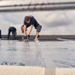 Two workers in protective gear are installing insulation panels on a roof. One person is drilling while the other is standing behind and working. Both are wearing knee pads and gloves. The scene is framed by the rungs of a ladder in the foreground.