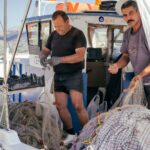 Two fishermen on a boat handle fishing nets filled with their catch. One man is in black clothing and gloves, while the other is in a purple shirt. The boat is docked and surrounded by mountains in the background. Various equipment is visible around them.