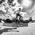 Black and white image of a shirtless skateboarder mid-air performing a trick at a skatepark. The skateboarder’s shadow is visible on the ground, and a clear sky with scattered clouds is in the background. Trees and other skaters can be seen on the sides.